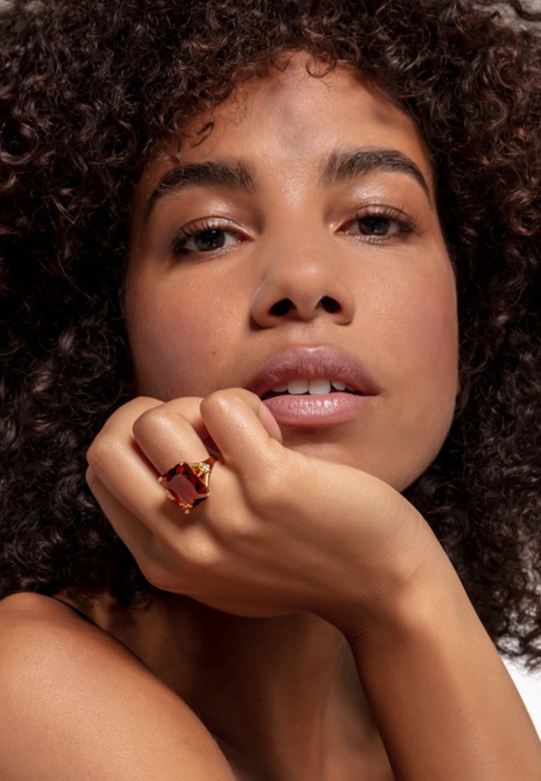 Bague en or ornée d'une grande pierre précieuse rectangulaire rouge. Monture courbée avec des accents texturés, portée sur une main près du visage.