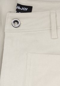 Beige katoenen stof met een diagonale weving, met een zilveren metalen knop en een gestikte zak. Zwarte label met "U&JOY" zichtbaar.