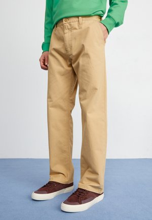 Beige cotton trousers with a relaxed fit, featuring a button closure and side pockets, paired with dark burgundy sneakers.