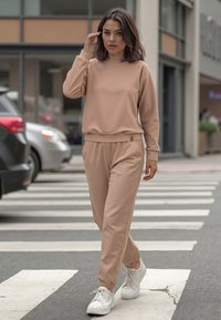 Púder bézs színű pulóver és jogger szett, puha anyagból készült. Bordás mandzsettákkal és diszkrét logós címkével rendelkezik. Fehér sportcipővel viselték.