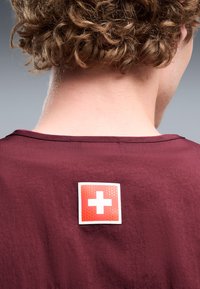 Personne aux cheveux bouclés portant une chemise grenat avec un écusson croix suisse rouge et blanche sur le haut du dos.