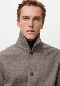 Abrigo de lana gris con cuello alto y cierre de botones. Presenta una tela texturizada y un diseño sencillo, combinado con un suéter de cuello alto negro debajo.
