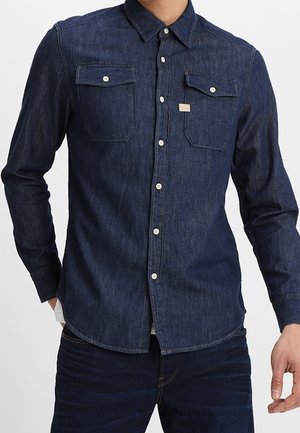 Donkerblauw spijkerblouse met lange mouwen, twee voorzakken, knoopsluiting en een klein merkpatch boven de linkerzak.