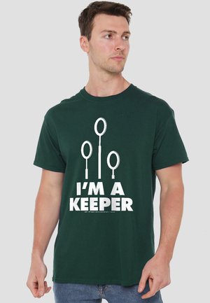 Man wearing dark green t-shirt with three white Quidditch goalposts and text "I'M A KEEPER" on front, looking to his right.