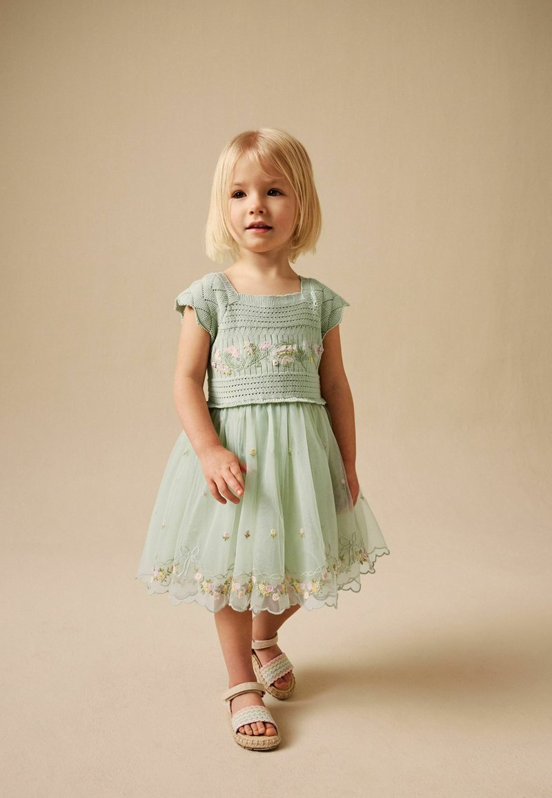Petite fille aux cheveux blonds portant une robe verte claire brodée et des sandales beige, debout devant un fond beige.
