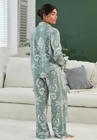 Vaaleanvihreä silkkinen pyjamapuku, jossa on valkoisia kukkakuoseja. Paidassa on kaulus; housut ovat leveälahkeiset, jotka täydentävät muotoilua.