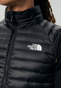 Crni puffer prsluk s visokim ovratnikom i zatvaranjem na patentni zatvarač, s bijelim logotipom s lijeve strane. Materijal ima glatku, sjajnu teksturu.