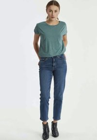 Camisa azul-petróleo de mangas curtas, combinada com jeans justos azuis e botas de tornozelo pretas. A camisa tem um decote redondo e um corte descontraído.