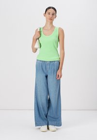 Top verde de canalé combinado con pantalones anchos de mezclilla azul sueltos. La modelo sostiene un bolso blanco. Conjunto simple y casual contra un fondo neutro.