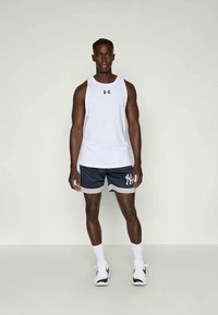 Atletski muškarac stoji, nosi bijelu majicu bez rukava Under Armour, tamnoplave kratke hlače New York Yankees, bijele čarape i bijele tenisice.