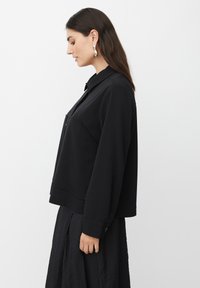 Vrouw met lang, donker haar draagt een zwarte kraagjasje met knoopmanchetten en een gestructureerde zwarte rok, poserend in profiel tegen een effen achtergrond.