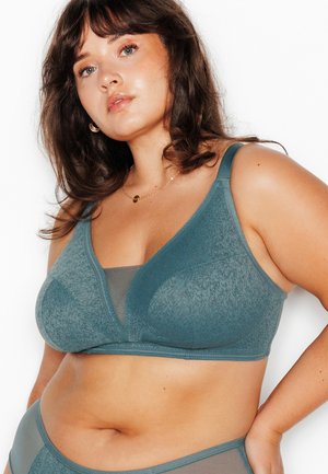 Donna con capelli castani fino alle spalle che indossa un reggiseno verde acqua a fantasia e mutandine a vita alta abbinati, in posa su sfondo bianco.
