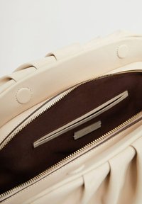 Sac fourre-tout beige avec une surface texturée, fermeture zippée et intérieur marron foncé. Équipé de quincaillerie dorée et d'une fente pour carte à l'intérieur.