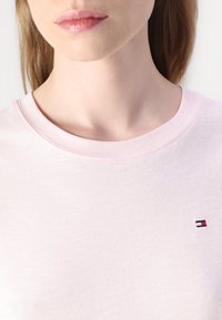 Tricou din bumbac roz deschis, cu guler rotund și mâneci scurte. Prezintă un mic logo brodat în roșu, alb și albastru pe piept.