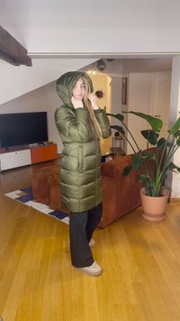 Maslinasto zelena jakna na preklop s kapuljačom, s patentnim zatvaračem i prošivenim dizajnom. Nošena s crnim hlačama i bež cipelama na drvenom podu.