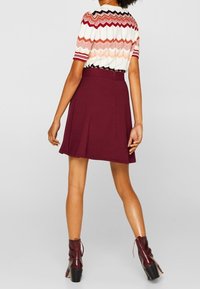 Rood-witte top met chevronpatroon en korte mouwen, gecombineerd met een bordeauxrode geplooide rok. Het model draagt glanzende bordeauxrode enkelboots.