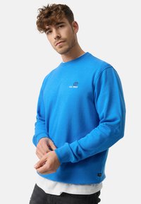 Sudadera azul de cuello redondo hecha de tela suave, con puños de canalé y un pequeño logotipo en el pecho. Llevada sobre una camisa blanca.