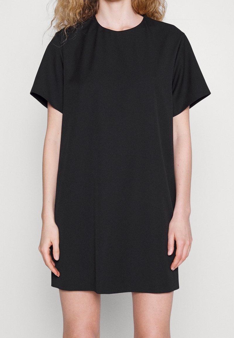 Femme aux cheveux bouclés portant une robe noire simple à manches courtes, debout devant un fond uni.