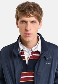 Marineblå jakke med stående krave, prydet med lapper og frontlynlås; lagdelt med en stribet polo t-shirt i burgunderrød, hvid og gul.