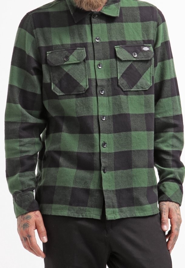 Homme portant une chemise à carreaux verte et noire à boutons avec deux poches poitrine et un pantalon noir.