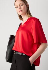 Blusa rossa di raso a maniche corte con scollatura a V, abbinata a pantaloni neri e a una borsa tote nera. Texture liscia e vestibilità morbida.