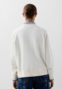Weißer Sweatshirt mit geripptem Saum und Bündchen, darunter sichtbar ein blauer Jeanshemd mit Kragen. Rückansicht zeigt eine glatte Stofftextur.