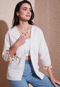 Kimono leggero di colore crema con maniche a tre quarti, presenta un tessuto strutturato e dettagli in frange sulle maniche. Abbinato a pantaloni blu.