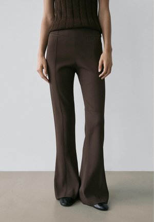 Slim-fit, high-waist brown pants with front seam details worn with black shoes and a sleeveless brown top against a plain background.