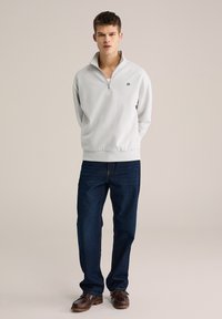 Hanorac gri deschis cu fermoar, realizat dintr-un material moale, cu un logo mic negru pe piept. Asortat cu blugi bleumarin închis și încălțăminte maro.