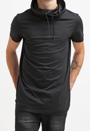 Mężczyzna w czarnej koszulce z krótkim rękawem i kapturem ze sznurkami, pokazujący tatuaż kompasu na lewym przedramieniu, stojący z rękami za plecami.