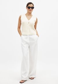 Cremfarbener, gerippter ärmelloser Oberteil mit V-Ausschnitt und Knöpfen, kombiniert mit weißen, weit geschnittenen Leinenhosen und braunen Flip-Flops. Modell trägt schwarze Sonnenbrille.