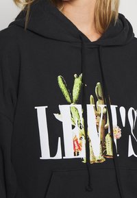 Haut à capuche noir avec un grand logo Levi's et des graphiques de cactus et de fleurs colorés. Comprend une capuche avec cordon de serrage et une poche kangourou.