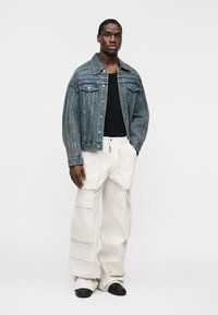 Άνδρας που στέκεται φορώντας μπλε τζιν μπουφάν, μαύρο πουκάμισο, φαρδιά λευκή παντελόνια cargo και μαύρα παπούτσια σε απλό λευκό φόντο.