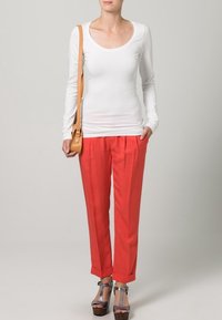 Witte lange mouwen top gecombineerd met oranje broek. De broek heeft een relaxte pasvorm en opgerolde zoom, aangevuld met metallic sandalen. Bruine handtas.