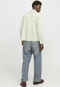 Camicia button-up beige chiaro con una vestibilità rilassata e maniche lunghe, abbinata a jeans svasati blu chiaro e scarpe marroni.