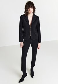 Una donna con capelli bagnati di lunghezza media indossa un blazer nero aderente con cerniera frontale, pantaloni neri slim e stivali neri con tacco a punta.