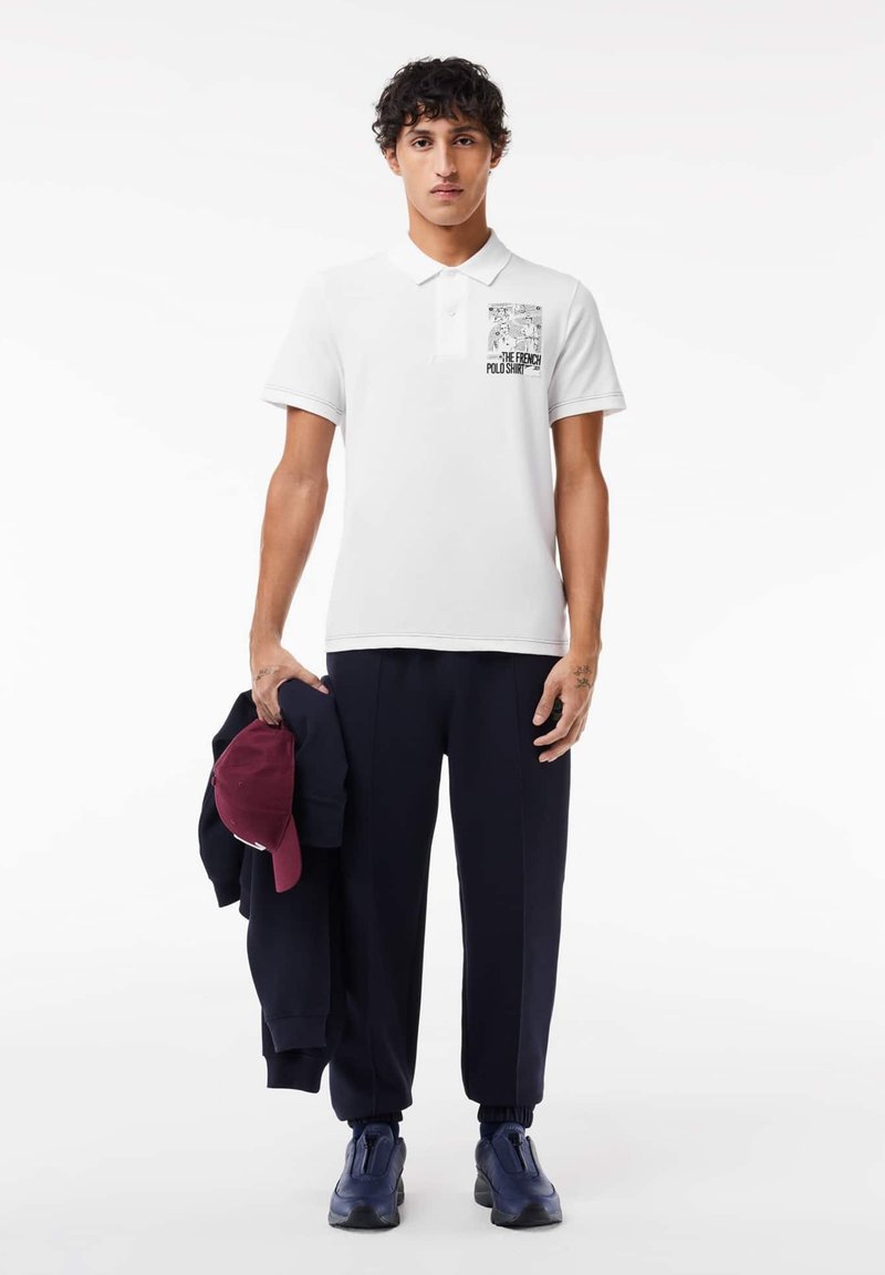 Chemise polo blanche avec un graphique noir sur la poitrine gauche, associée à un pantalon de jogging bleu marine et un sweat-shirt noir, tenant une casquette bordeaux.