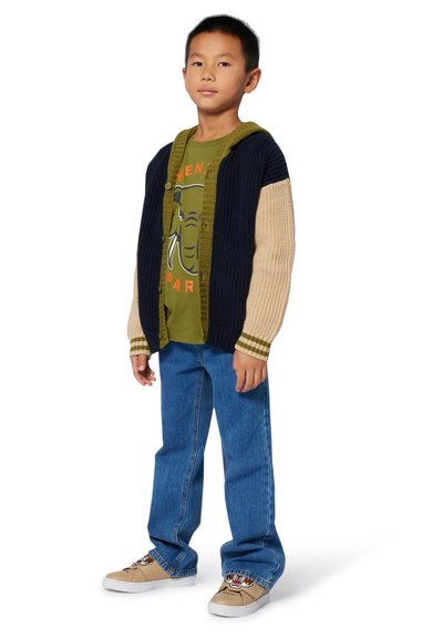 Cardigan boutonné en bleu marine et beige avec une texture côtelée ; T-shirt graphique vert ; jeans bleus ; chaussures crème avec un motif sur le bout.