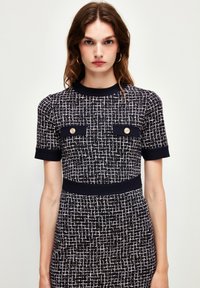 Vestido de punto con mangas cortas, que presenta un patrón texturizado en blanco y negro, botones decorativos dorados en los bolsillos de parche y una silueta ajustada.