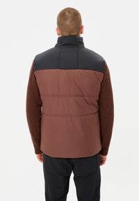 Mann mit kurzem Haar, der eine braune und schwarze gepolsterte Weste über einem braunen Sweater und schwarzen Hosen trägt, mit dem Rücken zu einer weißen Wand.