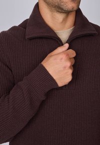 Pull en marron foncé avec une texture côtelée, un col zippé et des manches longues. Le tissu semble doux et chaud, adapté pour un usage décontracté.