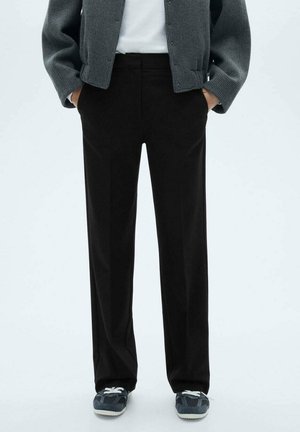 Osoba ubrana w czarne spodnie o prostych nogawkach, szary dziergany sweter, białą koszulę i ciemne trampki z białymi sznurówkami, stojąca z rękami w kieszeniach.