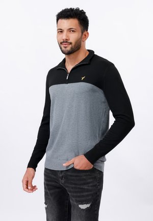 Sweatshirt à col noir et manches longues, présentant un corps gris et un emblème doré sur la poitrine. Fabriqué en tissu doux.