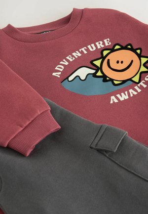 Bordeauxrotes Sweatshirt mit geripptem Rundhalsausschnitt, versehen mit einem Sonnenmotiv und dem Text "ABENTEUER ERWARTET." Graue Fleece-Hosen mit einer Seitentasche.