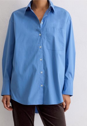 Osoba nosząca luźną, jasnoniebieską koszulę zapinaną na guziki z kieszenią na piersi oraz ciemne spodnie, stojąca na tle jednolitego białego tła.