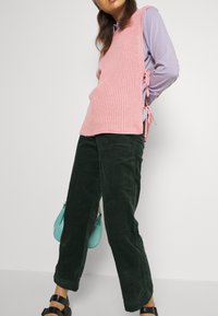 Rosa ribbstickad väst med sidoknytningar, lagd över en ljuslila skjorta, ihop med mörkgröna kordbyxor och en teal handväska.