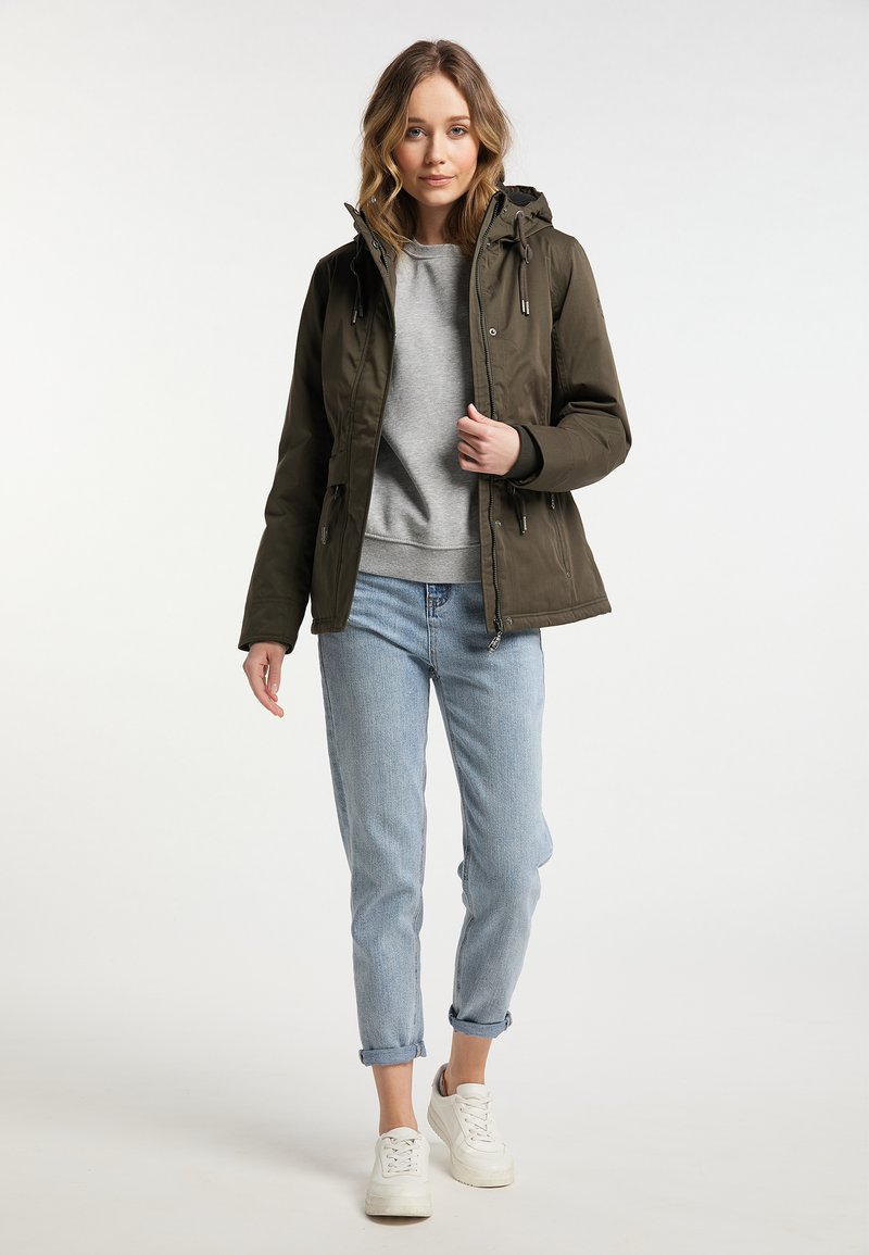 Dunkelgrüne Kapuzenjacke mit Reißverschluss, kombiniert mit einem grauen Sweatshirt, hellblauen Jeans und weißen Sneakern, vor einem neutralen Hintergrund.