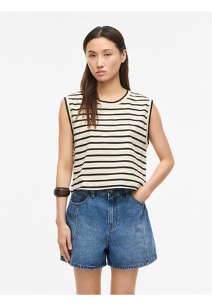 Femme portant un haut sans manches rayé noir et blanc, un short en jean bleu taille haute et un grand bracelet sombre à son poignet droit.