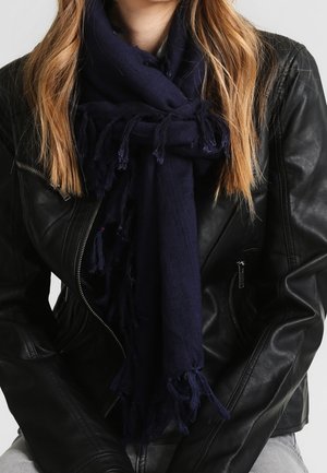 Personne portant une écharpe à franges bleu foncé nouée et une veste en cuir noire, avec de longs cheveux châtain clair visibles sur les épaules.