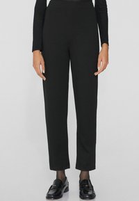 Pantalon noir, ajusté, en tissu lisse avec une taille élastique. Associé à des mocassins noirs, les mains reposent sur les côtés.